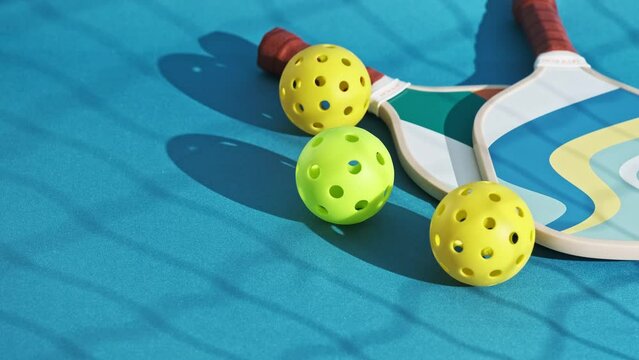 three pickleball balls rolling on the floor of a blue court that end up touching some paddles on the ground