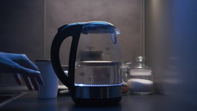 beautiful young female hand turns on a transparent glass electric kettle with water presses the button blue lamp lights up at dusk