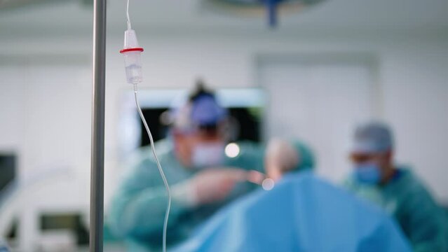 Saline solution dripping inside the drop counter. Surgeon carries out the operation at backdrop in blur.