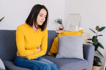 Young woman suffering stomach ache or abdominal pain while sitting on couch at home. Menstruation or pregnancy health care effects