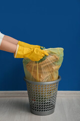 Female hands in rubber gloves pulling full garbage bag from trash bin near blue wall