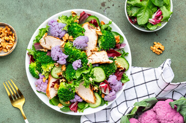 Healthy fresh salad from purple cauliflower, broccoli, grilled chicken breast, cucumbers, red onion, radicchio, spinach with walnuts, green table background, top view
