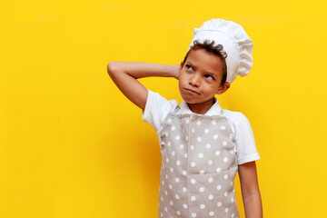 puzzled african american boy in chef's uniform and hat thinks and scratches his head on yellow...