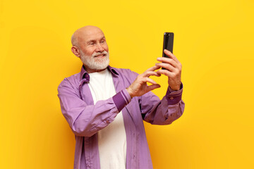 old bald grandfather with gray beard uses smartphone and selects online on yellow isolated background