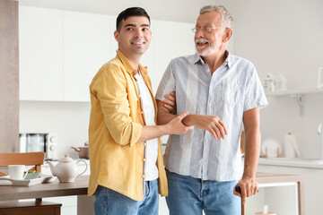 Young man helping his father with stick to walk in kitchen