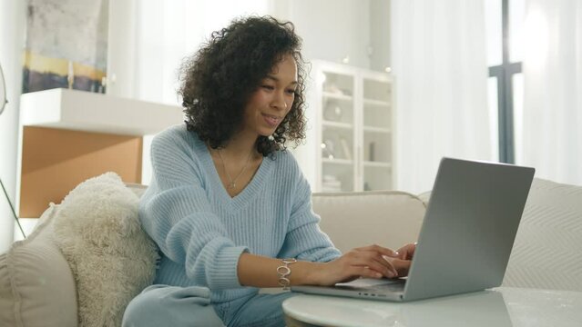 Portrait of beautiful stylish authentic female with afro hairstyle typing on laptop computer home. Casual woman of color having fun browsing Internet and checking videos on Social Media in living room