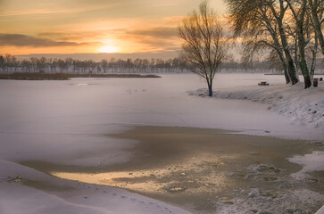 Snowy winter landscape with sunset in the background. The frozen Tisza backwater can be seen in Tiszalök, Hungary. Bare trees and branches are still visible in the picture. Hungarian rural landscape. 
