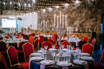 Close-up photo of chromium candlestick with five unlit white wax candles placed on the table prepared for wedding reception. Round tables with red chairs
