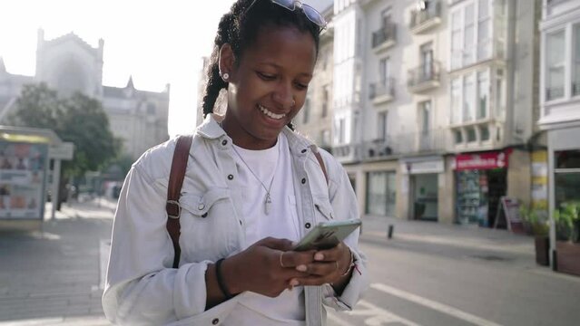 Beautiful young black woman walking down street with mobile in her hands, she looks smiling and funny at the phone screen, chatting and sends voice audios strolling through city.