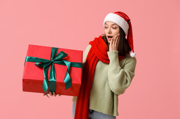 Shocked young woman in Santa hat with gift box on pink background