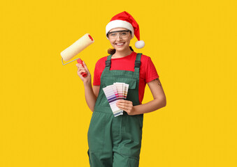Beautiful female painter in Santa hat with roller and color palettes on yellow background