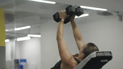 Woman lying on a bench in the gym doing an exercise for chest muscles. She spreads her arms to the sides with dumbbells.