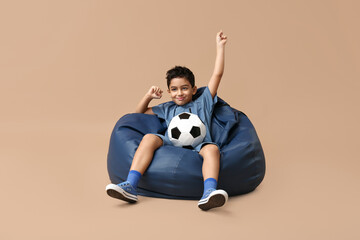 Happy little boy with soccer ball sitting in beanbag chair on beige background