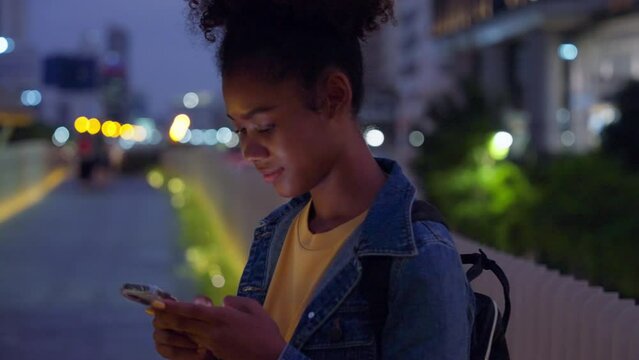 Beautiful young african woman standing in city street at night using mobile smartphone. Happy afro woman typing messages on her mobile phone