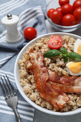 Tasty boiled oatmeal with egg, bacon and tomatoes served on light grey table, closeup