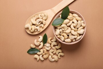 Tasty cashew nuts and green leaves on pale brown background, flat lay