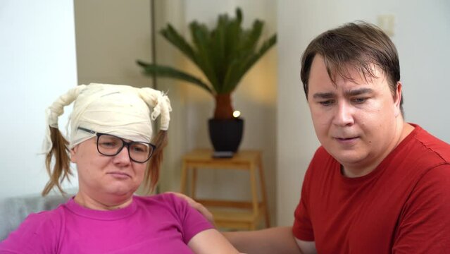 Woman with bandaged head, glasses playsfully pouts her lips, is capricious, incites her henpecked husband manipulates man patient with injury at home rehabilitation complains, helplessly asks for help