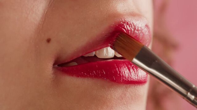 Young woman make-Up with a Red Lipstick on her lips close-up. Shooting of woman applying red lipstick. Make up routine, pink background