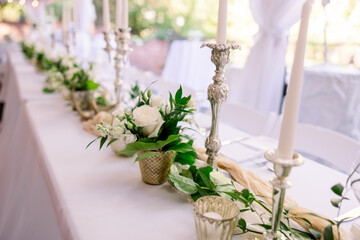 A gorgeous decorated head table at an outdoor wedding reception with candles, candelabra, roses, and greenery
