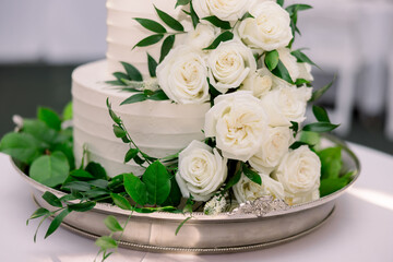 A close-up of a wedding cake at a wedding reception, decorated with lots of white roses and green leaves, on a silver platter