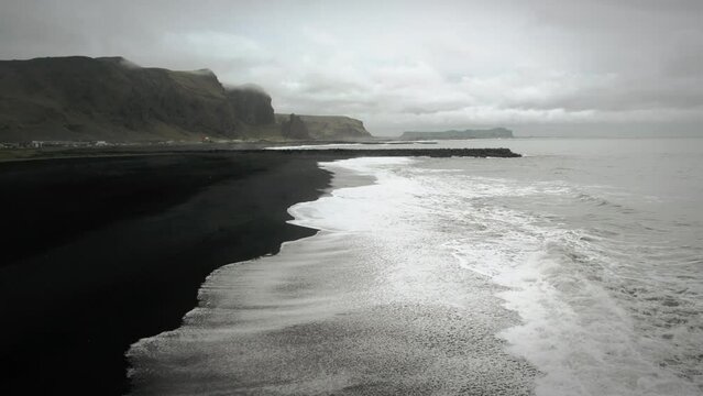 Cinematic aerial flying over black sand beach ocean scenery, moody dark scenery, town Vik, Iceland