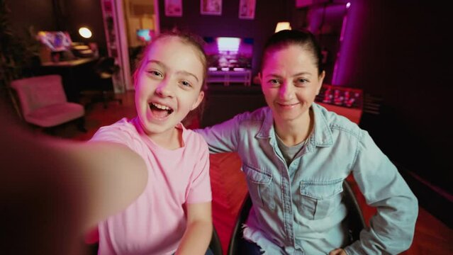 Cute kid teaching her mother how to do viral online videos for social media, filming in POV style. Mom learning how to do trending clips from influencer daughter in home illuminated by pink neon