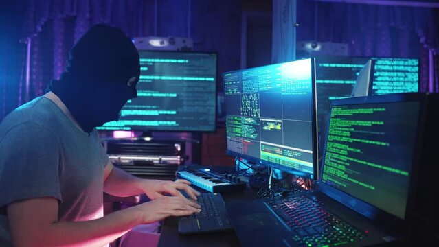 Hacker coding and listening music on programming computer at table. Male anonymous individual looking at notebook and hacking pc in neon color light. IT cybercrime of thief mask hacks virtual database