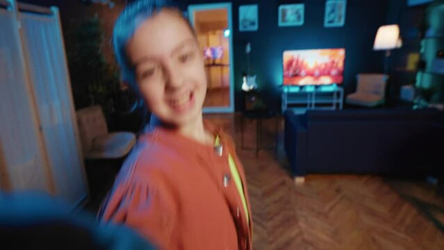 Kid films POV style dance tutorial video with smartphone in blue neon lit living room. Young dancer does viral online social media dancing challenge, capturing footage with selfie cellphone camera