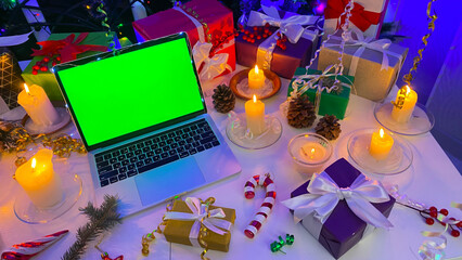 An open laptop with a chroma key on the screen lies on a table decorated with New Year's gifts, burning candles, cones, confetti, fir branches and striped caramel