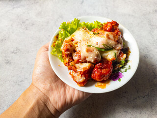 Man hand holding a plate of Korean style fried chicken with spicy sauce and mozzarella cheese sprinkled with scallion and sesame seed and lettuce on the side on a white plate.