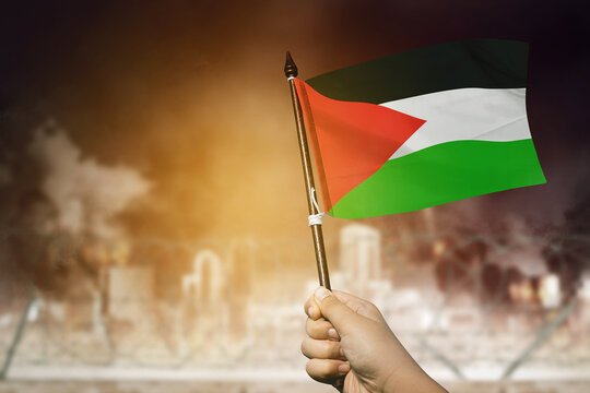 Closeup of a man holds a Palestinian flag in front of a burning city