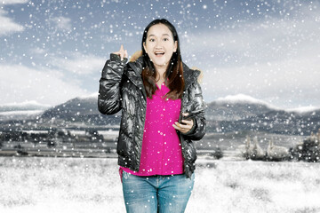 Portrait of a cheerful casual girl wearing winter jacket holding mobile phone and pointing finger away