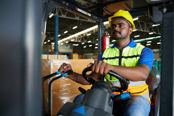 Indian worker driving a forklift and looking to something in warehouse storage