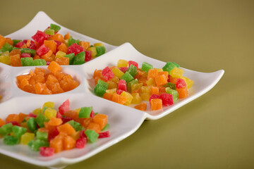 Sweet dessert in form candied fruit cubes close-up in a white menager on a yellow background. On yellow background, close-up dessert of colored candied fruit is laid out on white square-shaped plate. 
