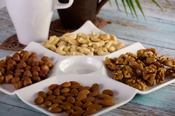 Assorted dried nuts of various types are laid out in close-up on a white square plate. A mix of hazelnuts, almonds, cashews and walnuts is laid out in a white bowl standing on a wooden table