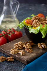 Walnut halves are laid out on wooden board on table next to plate of fresh chopped vegetables from cherry tomatoes and lettuce. Close up of beautiful original serving vegetable salad with walnuts