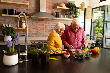 Happy caucasian senior couple preparing chopped vegetables in sunny kitchen, copy space