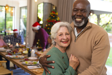 Two happy diverse male and female senior friends embracing, at christmas meal with friends