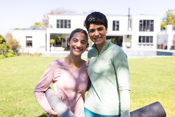 Portrait of happy biracial lesbian couple holding yoga mats embracing in sunny garden