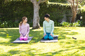 Happy biracial lesbian couple practicing yoga kneeling on mats in sunny garden, copy space