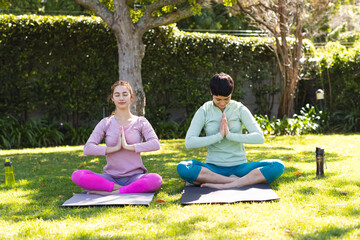 Happy biracial lesbian couple practicing yoga meditation sitting on mats in sunny garden, copy space