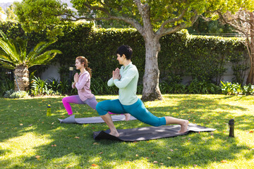 Happy biracial lesbian couple practicing yoga standing in lunge pose in sunny garden, copy space