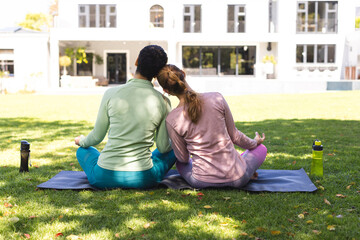 Rear view of biracial lesbian couple sitting in yoga meditation touching heads in sunny garden