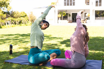 Happy biracial lesbian couple practicing yoga sitting stretching in sunny garden