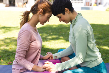 Happy biracial lesbian couple practicing yoga sitting holding hands and touching heads in garden