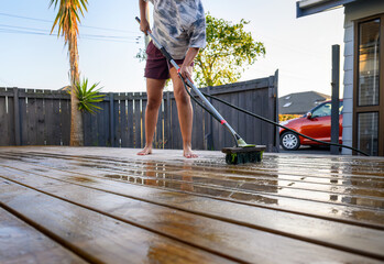 Woman cleaning wooden deck with brush and water sprayer. Red car parked outside the house. Auckland.