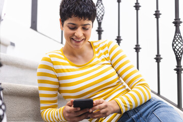 Happy biracial woman using smartphone sitting on stairs at home - Powered by Adobe