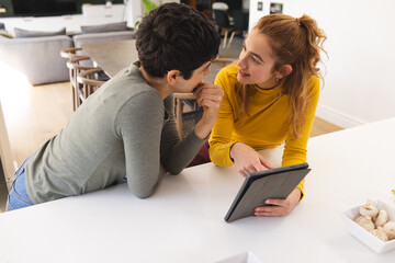 Happy biracial lesbian couple using tablet, standing leaning on countertop in sunny kitchen