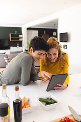 Happy biracial lesbian couple using tablet, leaning on countertop in sunny kitchen, copy space