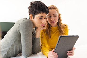 Focused biracial lesbian couple using tablet, leaning on countertop in sunny kitchen
